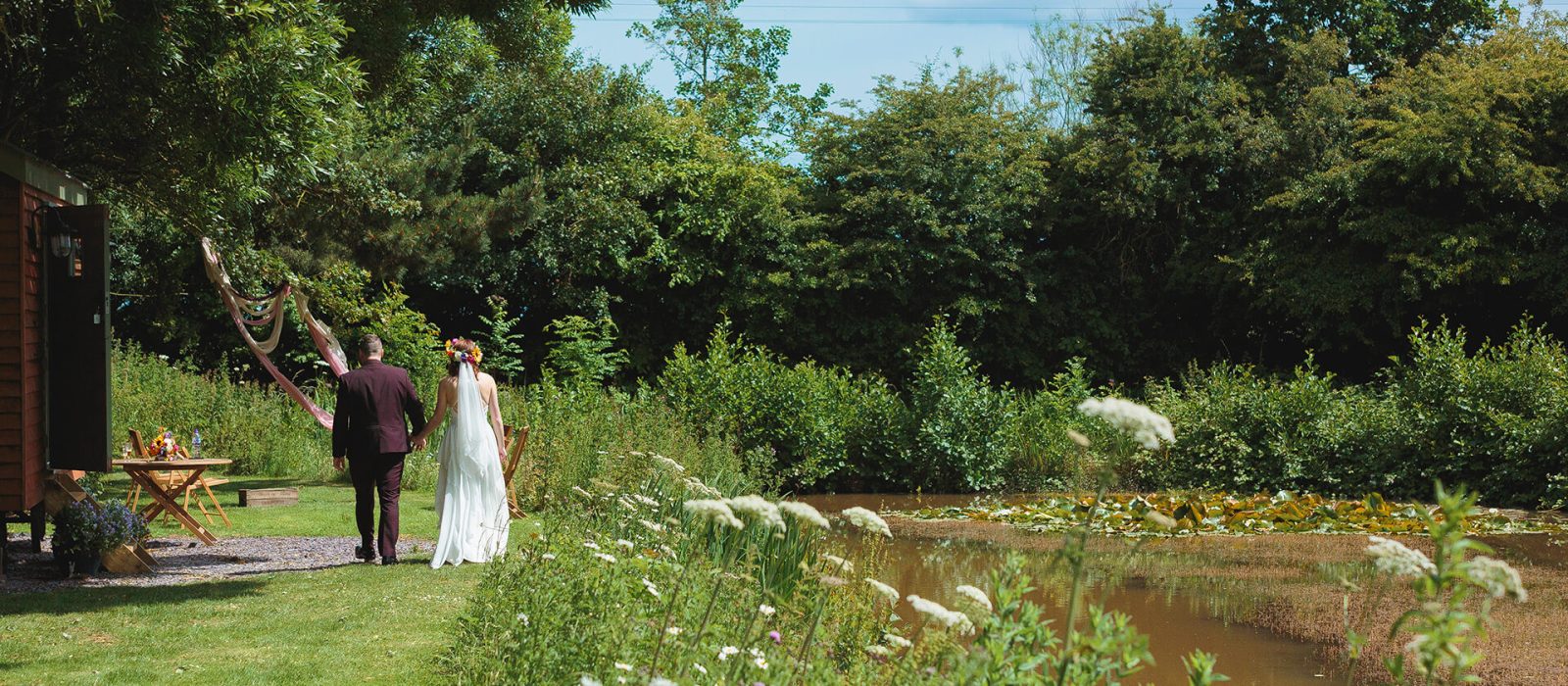 A bride in a white gown and floral crown and a groom in a burgundy suit walk hand-in-hand past a rustic shepherd's hut and a blooming lily pond, with wildflowers and tall grasses in the foreground, at a Yorkshire wedding venue. Featured by Northern Wedding Films for York Wedding Network.