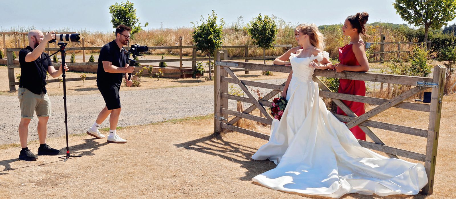 Wedding videographers Mike and Nathan behind the scenes filming a bride in an off-the-shoulder gown with bridesmaids at a rustic wooden gate at The Beverley Barn.
